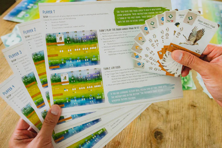 Wingspan board game player sheets and cards in hands, featuring bird illustrations and gameplay instructions.